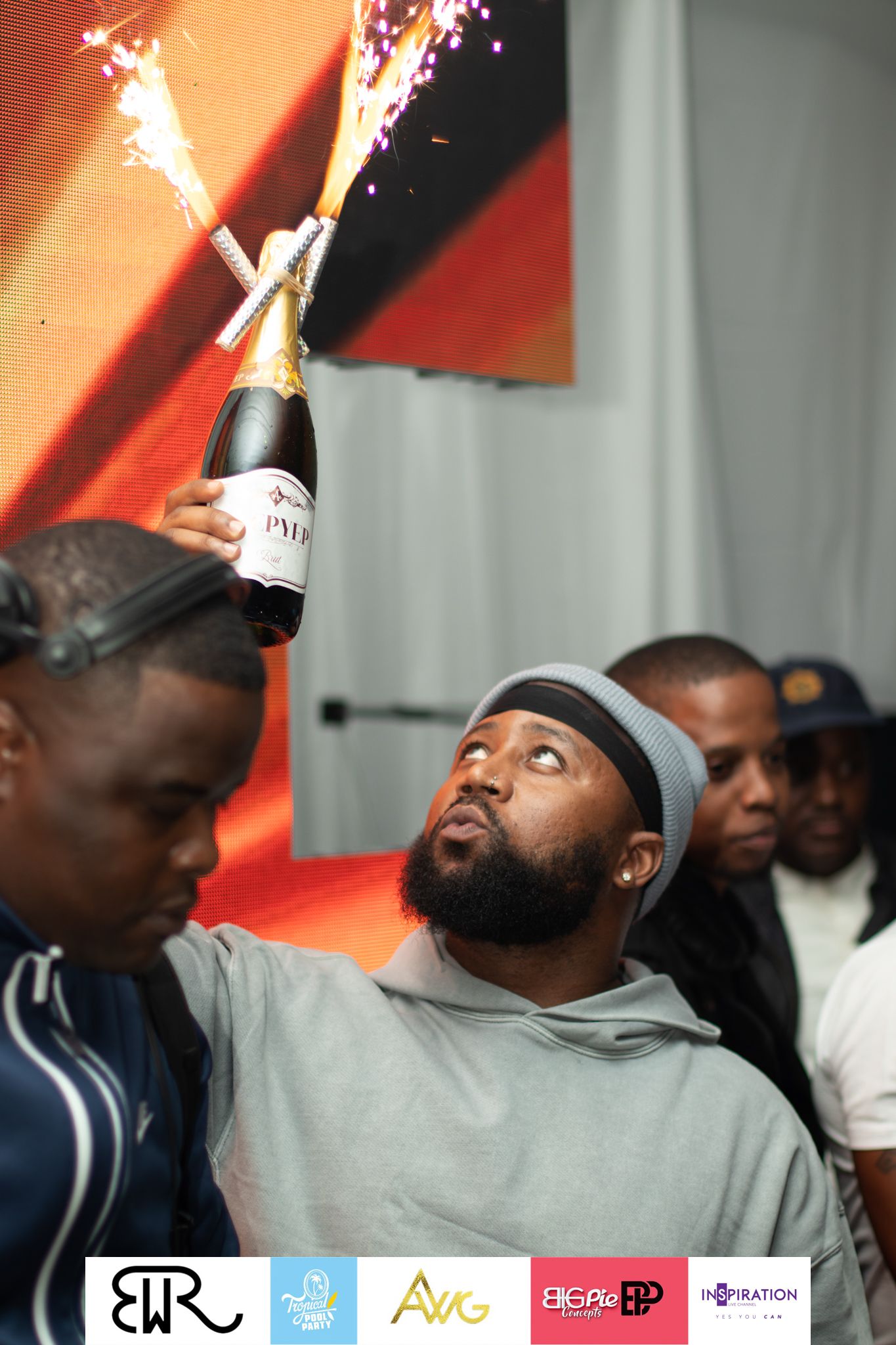 A celebratory moment as champagne sprays from a bottle at a festive party event with a red backdrop.
