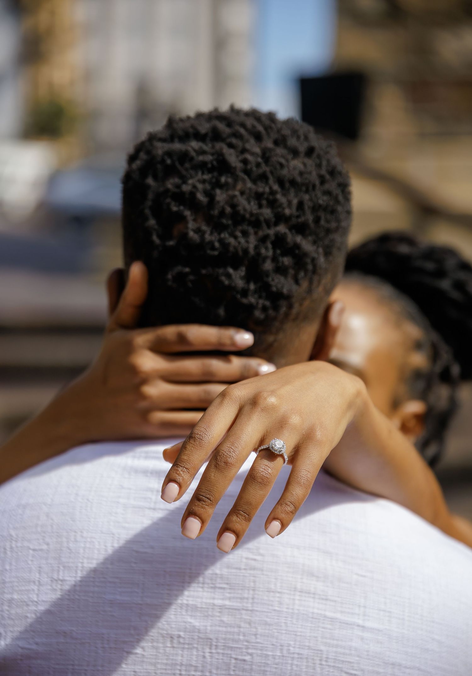 Hand with engagement ring rests lovingly on white-clothed shoulder with short curly dark hair in soft intimate moment.