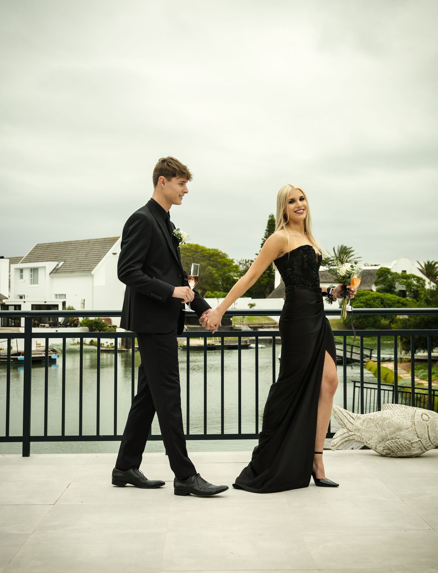 Elegant couple in formal black attire poses on a waterfront balcony with coastal homes in background.