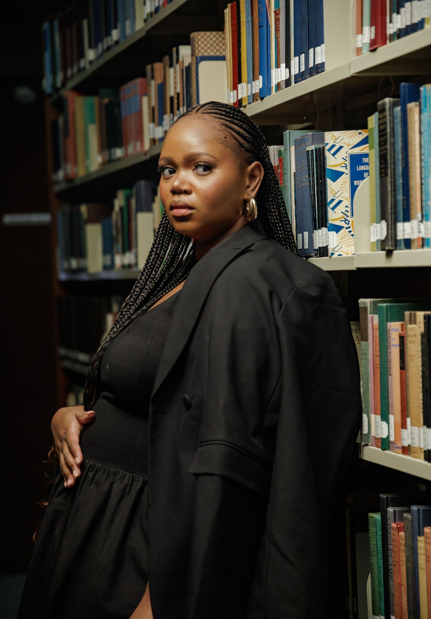 Professional portrait in a library wearing a black blazer and dress, standing between shelves of books with elegant lighting.