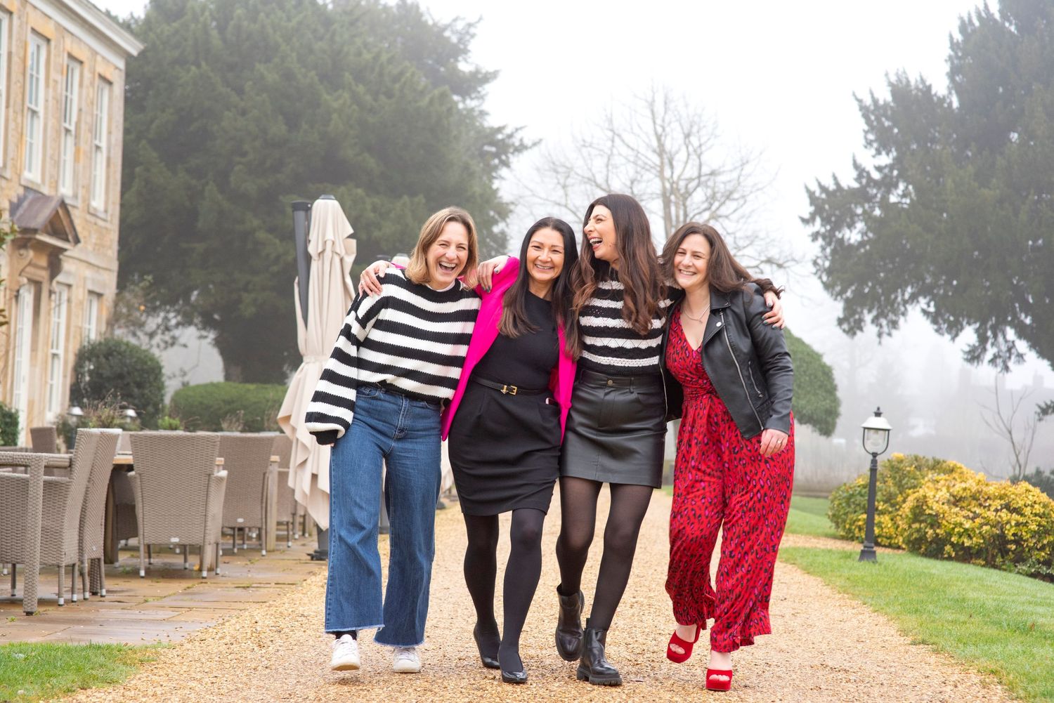A group walks arm-in-arm along a garden path at a stately venue wearing casual winter outfits in stripes and bright colors.