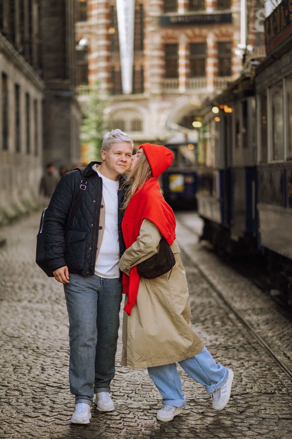 A couple shares playful moments on a cobblestone street wearing casual winter outfits.