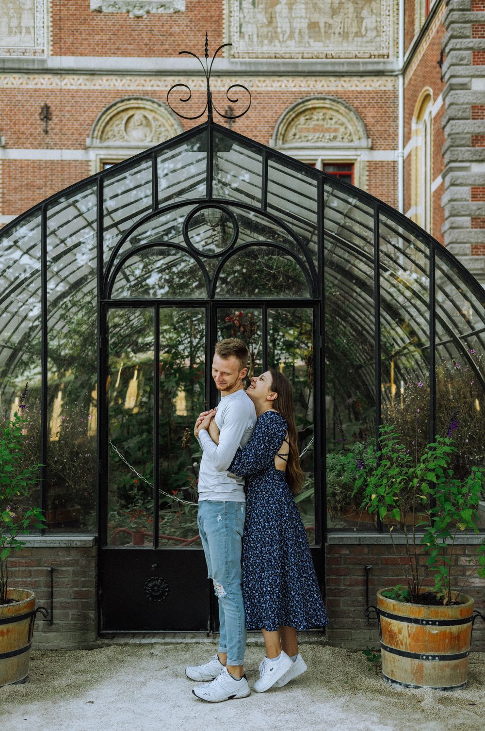 Two people embrace near a greenhouse with ornate iron architecture and brick walls.