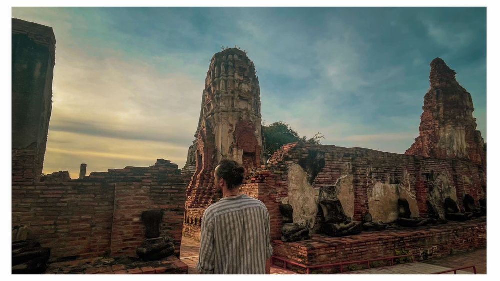 Someone observing ancient temple ruins at sunset in Ayutthaya, Thailand with crumbling stone structures and towers.