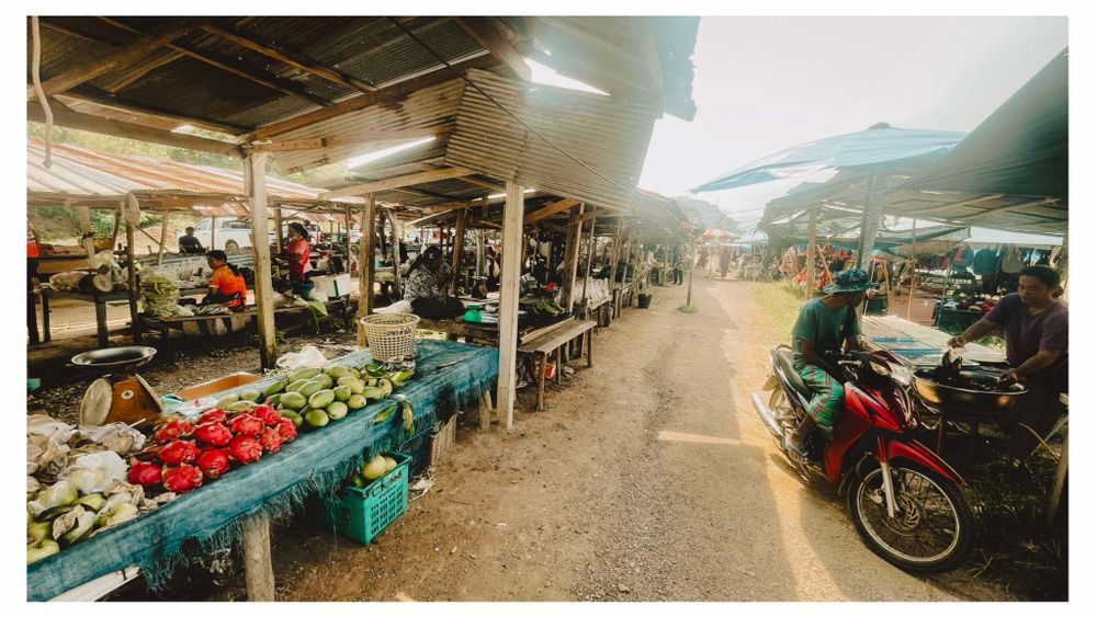View down a covered market aisle with stalls selling produce and goods, with motorcycles parked along the walkway.