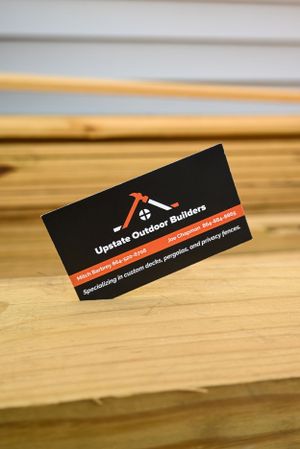 A black business card for Upstate Outdoor Builders featuring a crossed hammer and ruler logo on a wooden surface.