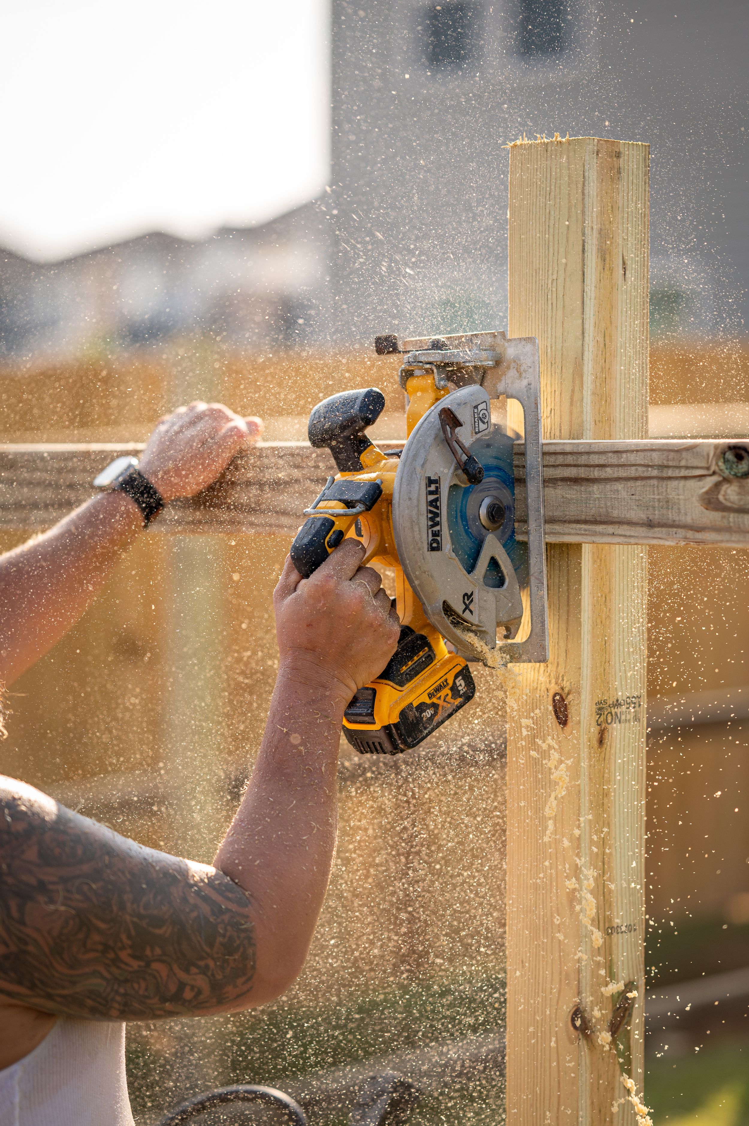 A close-up view of hands using a yellow circular saw to cut through wooden lumber with sawdust flying in sunlight.