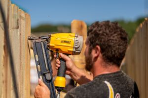 Someone using a yellow DeWalt nail gun to install wooden fence panels outdoors on a sunny day.