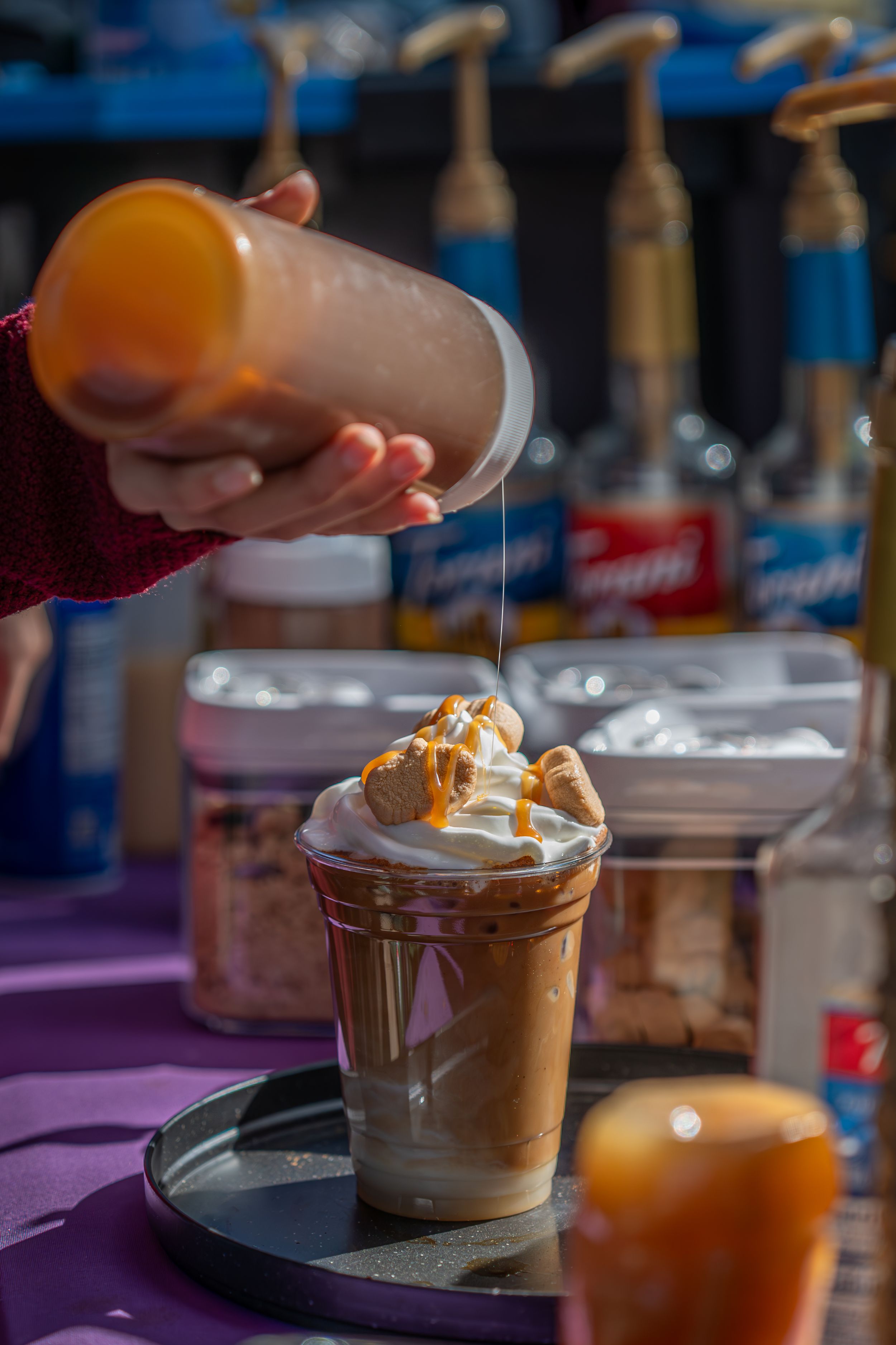 Someone pours caramel sauce over a frothy coffee drink topped with whipped cream, with blurred bottles in the background.