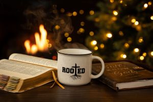 White ceramic mug with Rooted logo sits beside open books with Christmas lights and fireplace glow in background.