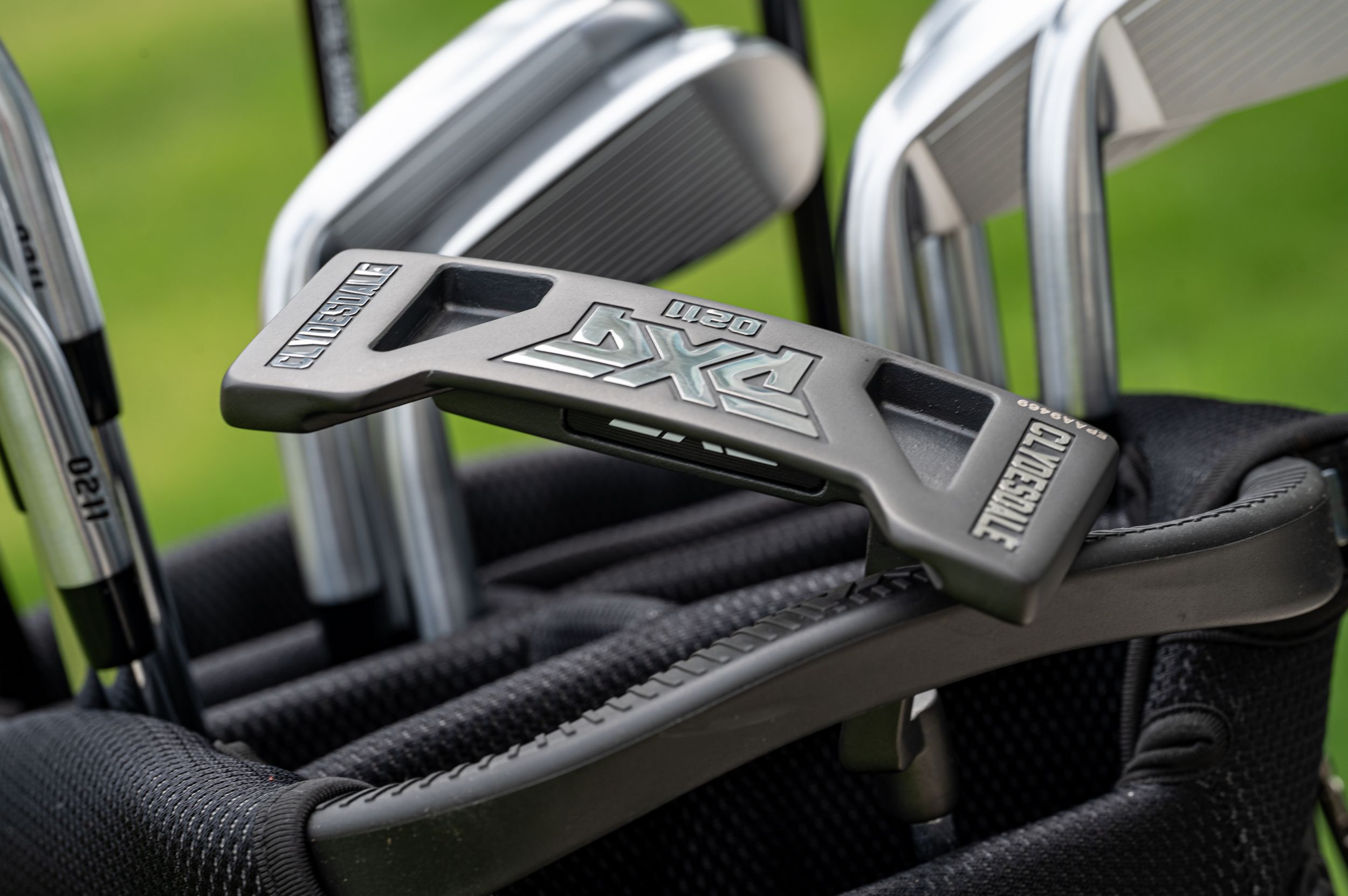 Close-up view of golf club heads showing Cobra RXH irons in a black golf bag on green grass background.