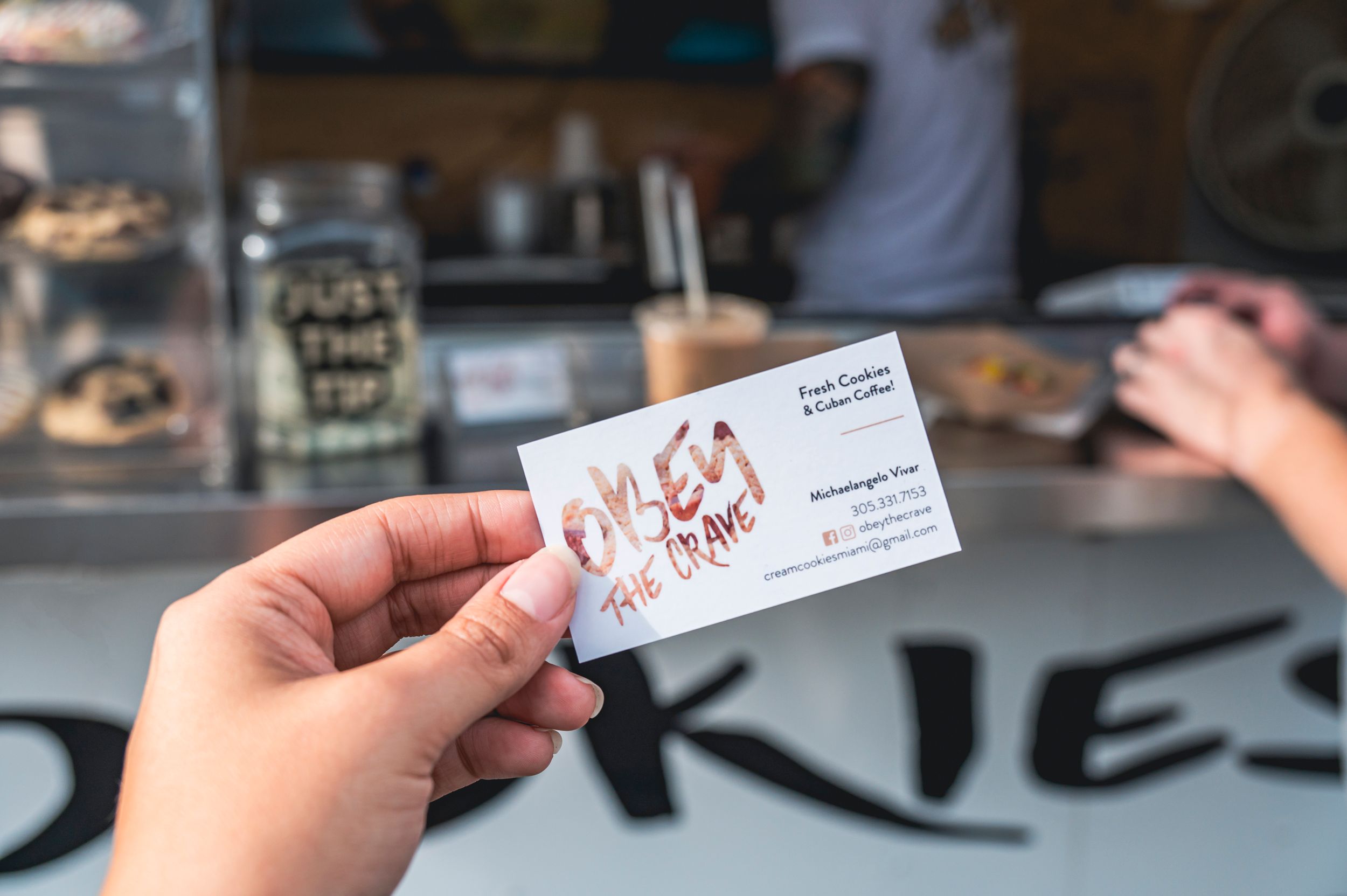 A hand holds a white business card in front of a blurry counter or retail display.