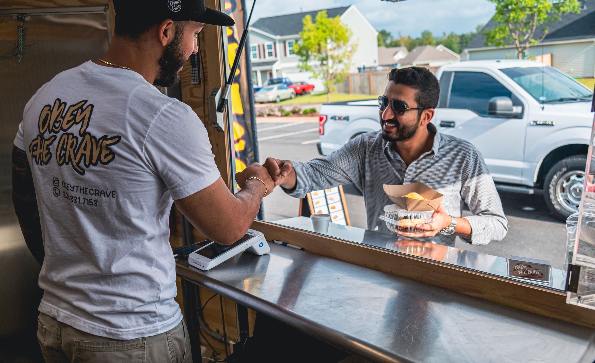 Two people in sunglasses chat and smile while seated at an outdoor cafe counter.