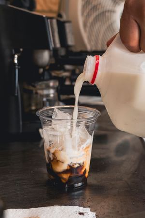 White milk being poured into a clear plastic cup containing iced coffee and cream.