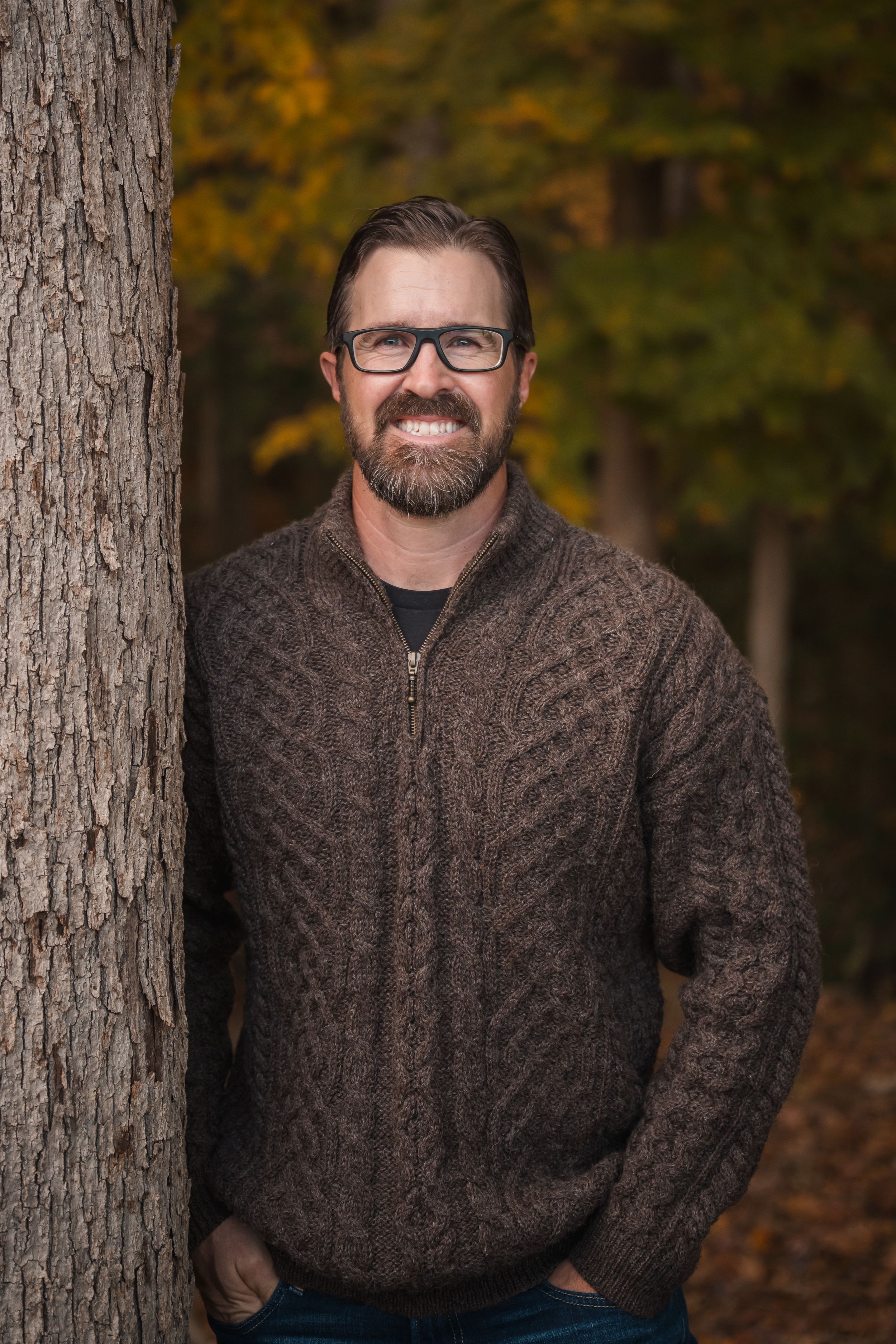 A person wearing glasses and a brown cable knit sweater poses outdoors against an autumn forest backdrop.