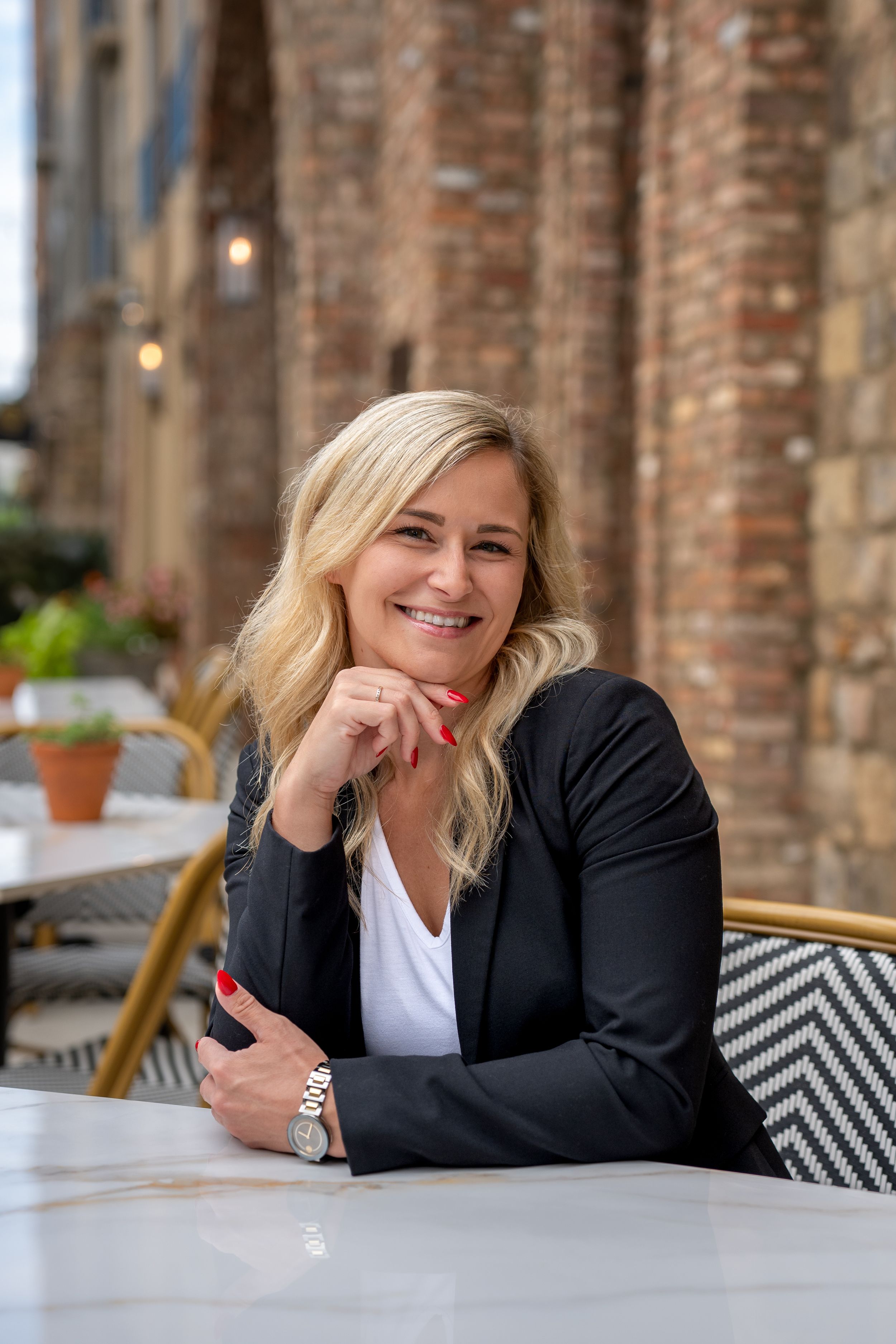 A person in a black blazer sits at an outdoor cafe table with patterned cushions.