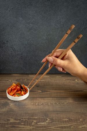 Hand using wooden chopsticks to pick up kimchi from a small white bowl on a rustic wooden table.