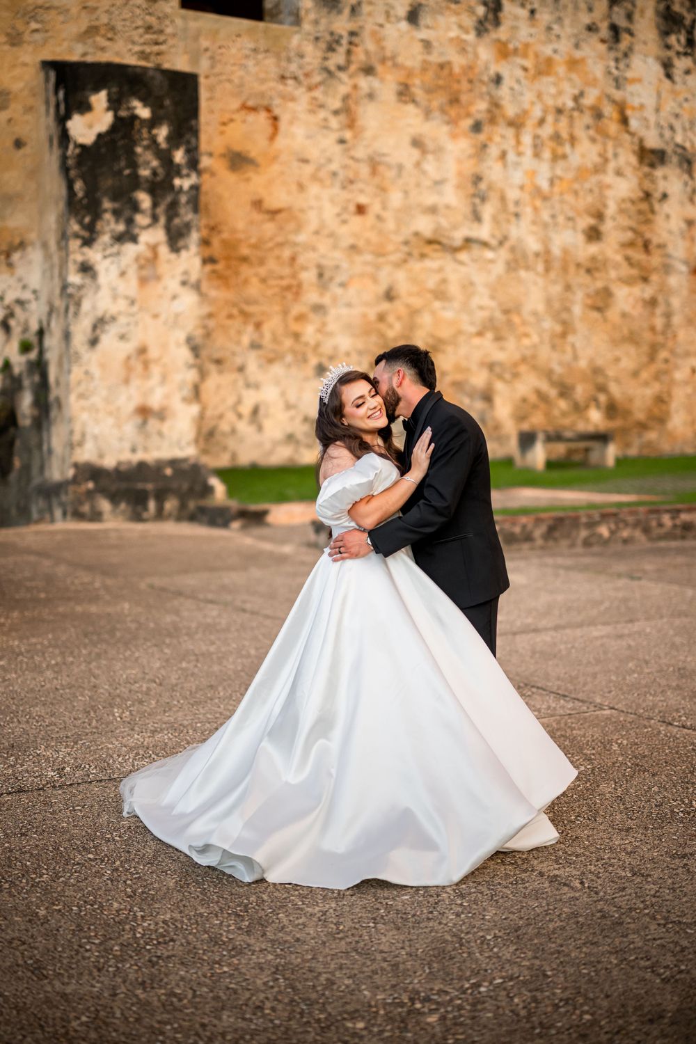 A newlywed couple embraces in front of a weathered fortress wall during their wedding photoshoot.
