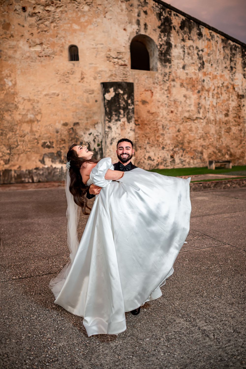 A groom lifts his bride in a flowing white dress against the backdrop of a historic stone building.