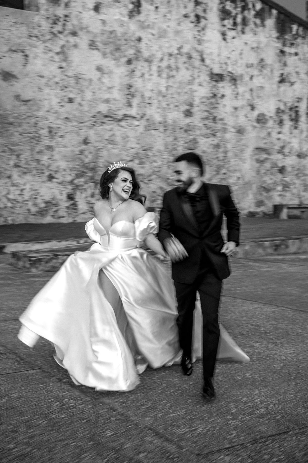 A joyful couple runs together along a stone wall in black and white wedding photography.