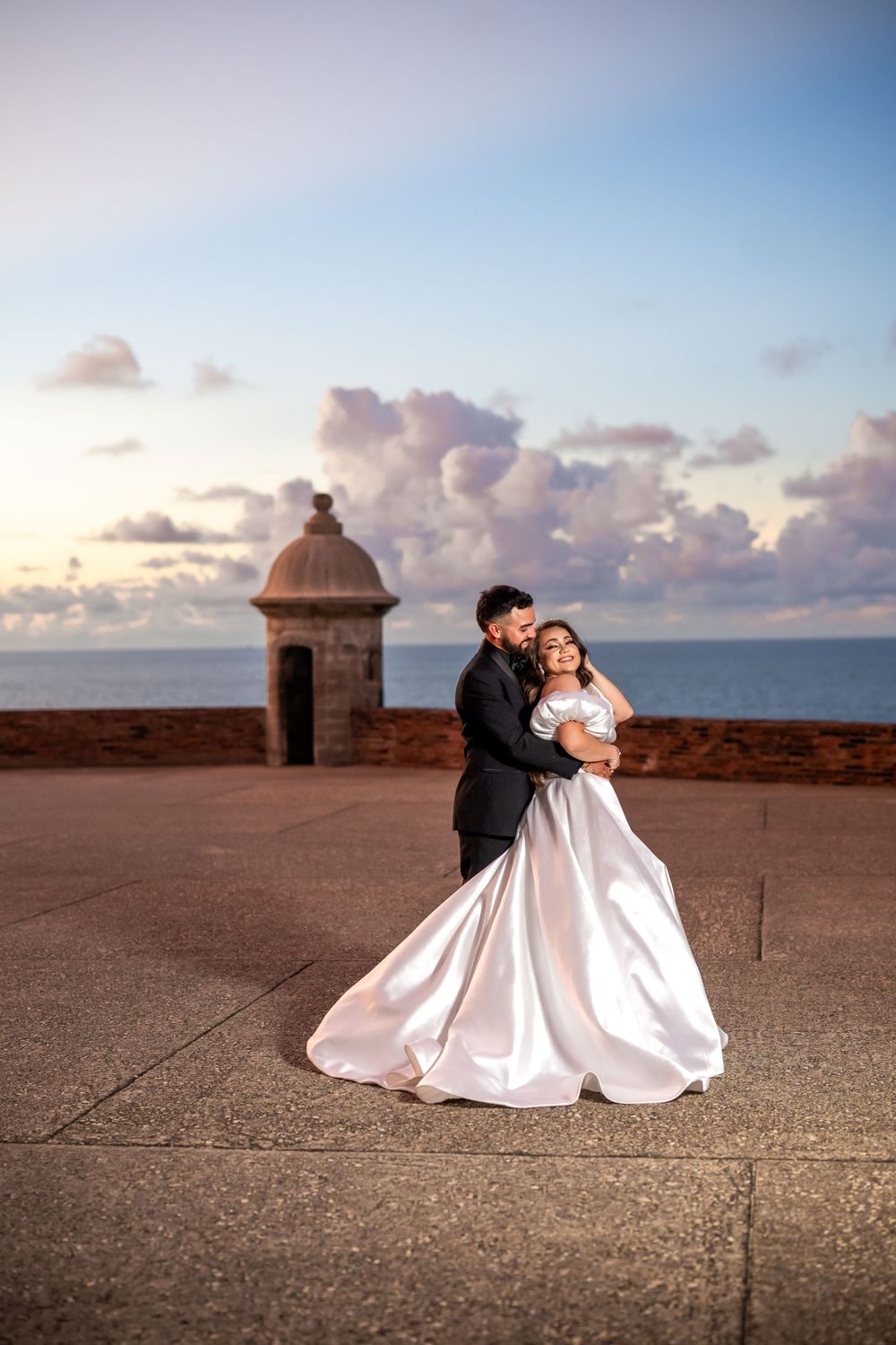 A couple embraces near a historic coastal watchtower during sunset.