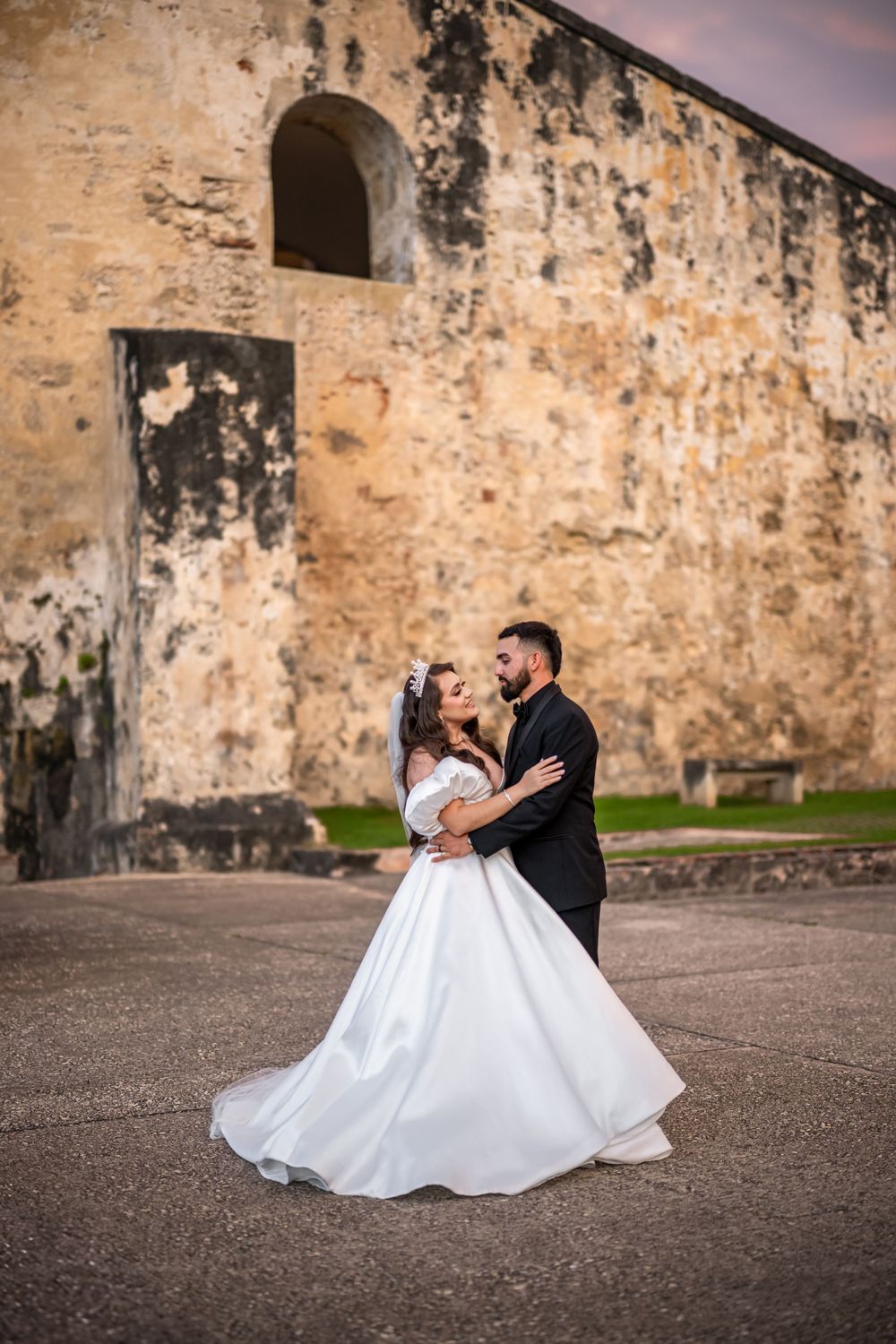 A couple shares a romantic moment in front of a historic stone fortress wall at sunset.