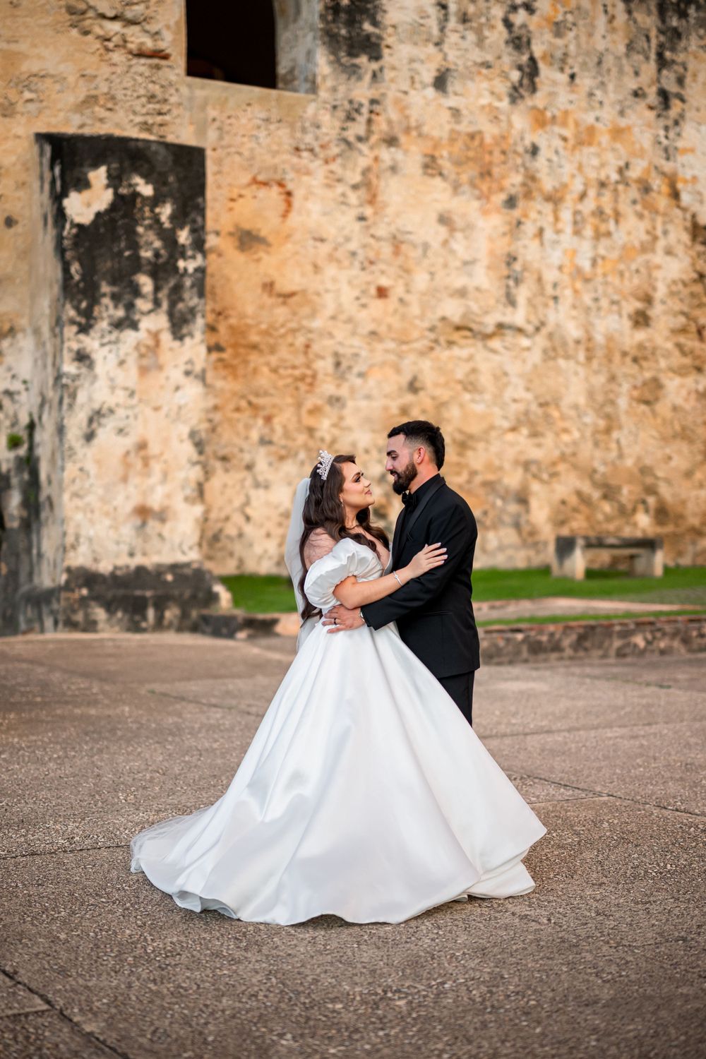 A bride in a flowing white gown dances with her groom against the backdrop of an ancient stone wall.