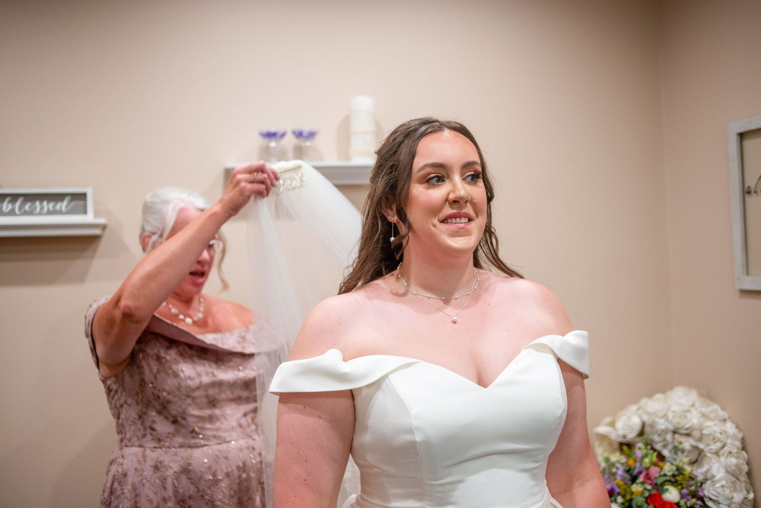 A bride in a white off-shoulder gown smiles while getting ready for her wedding.