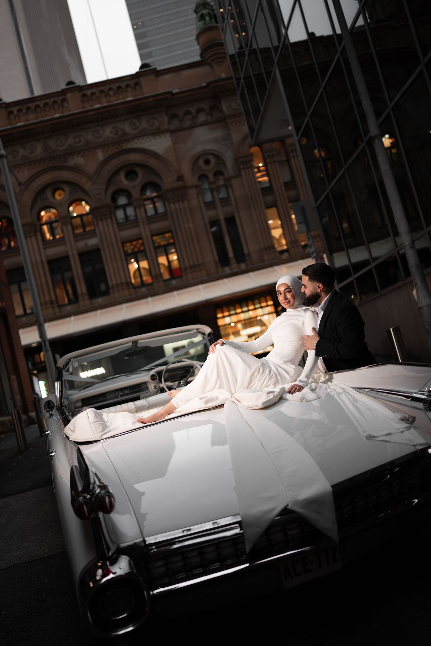 Bride in white dress reclining on vintage white convertible with groom in urban city setting.