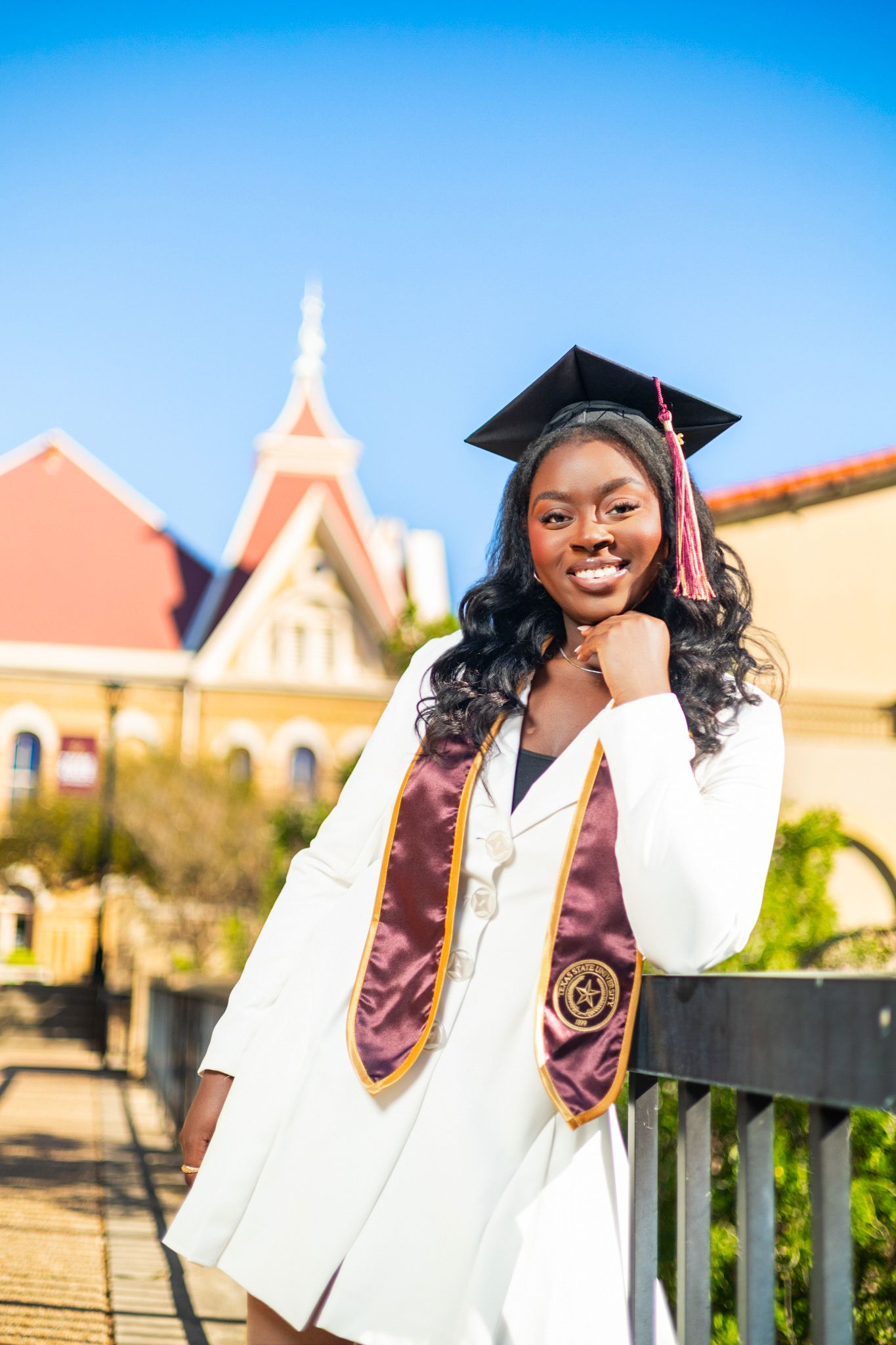 Graduate posing outdoors in white academic regalia with purple stole against Spanish-style architecture and blue sky.