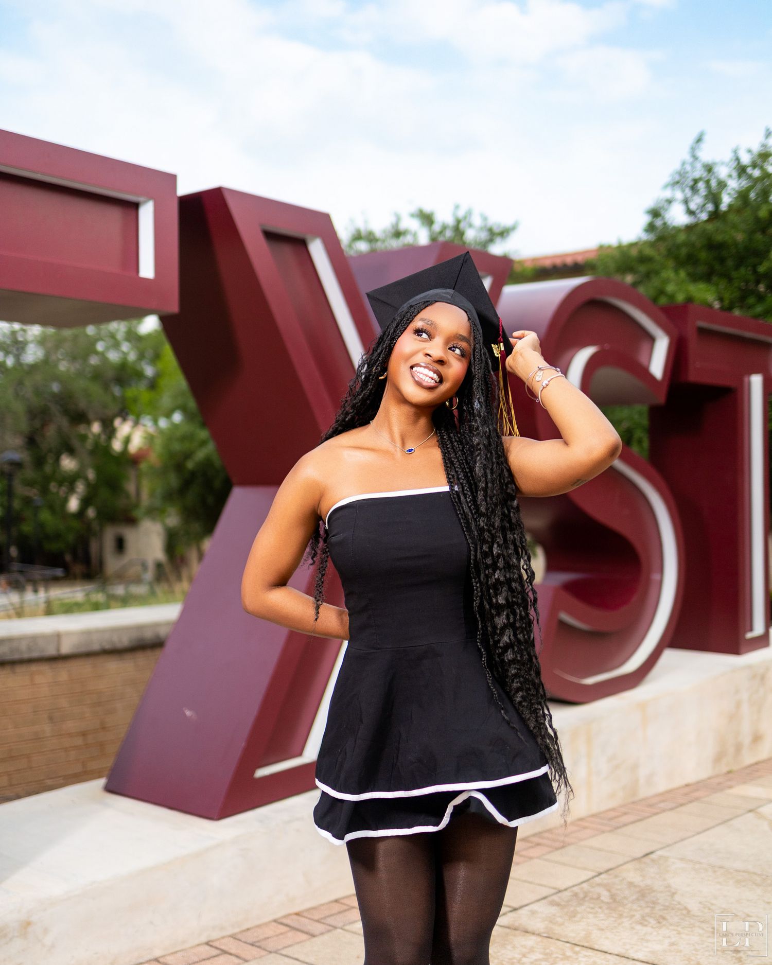 Joyful graduate in black dress poses in front of red architectural structure during photo session.