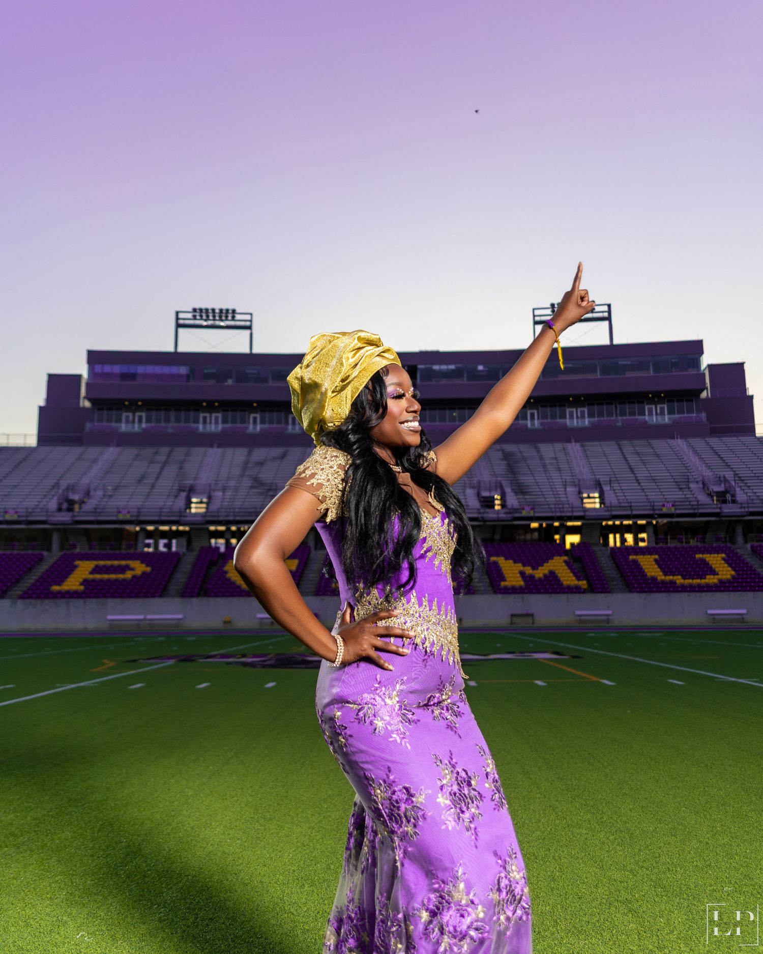 Graduate in sparkly purple dress celebrates on football field with arms raised triumphantly.