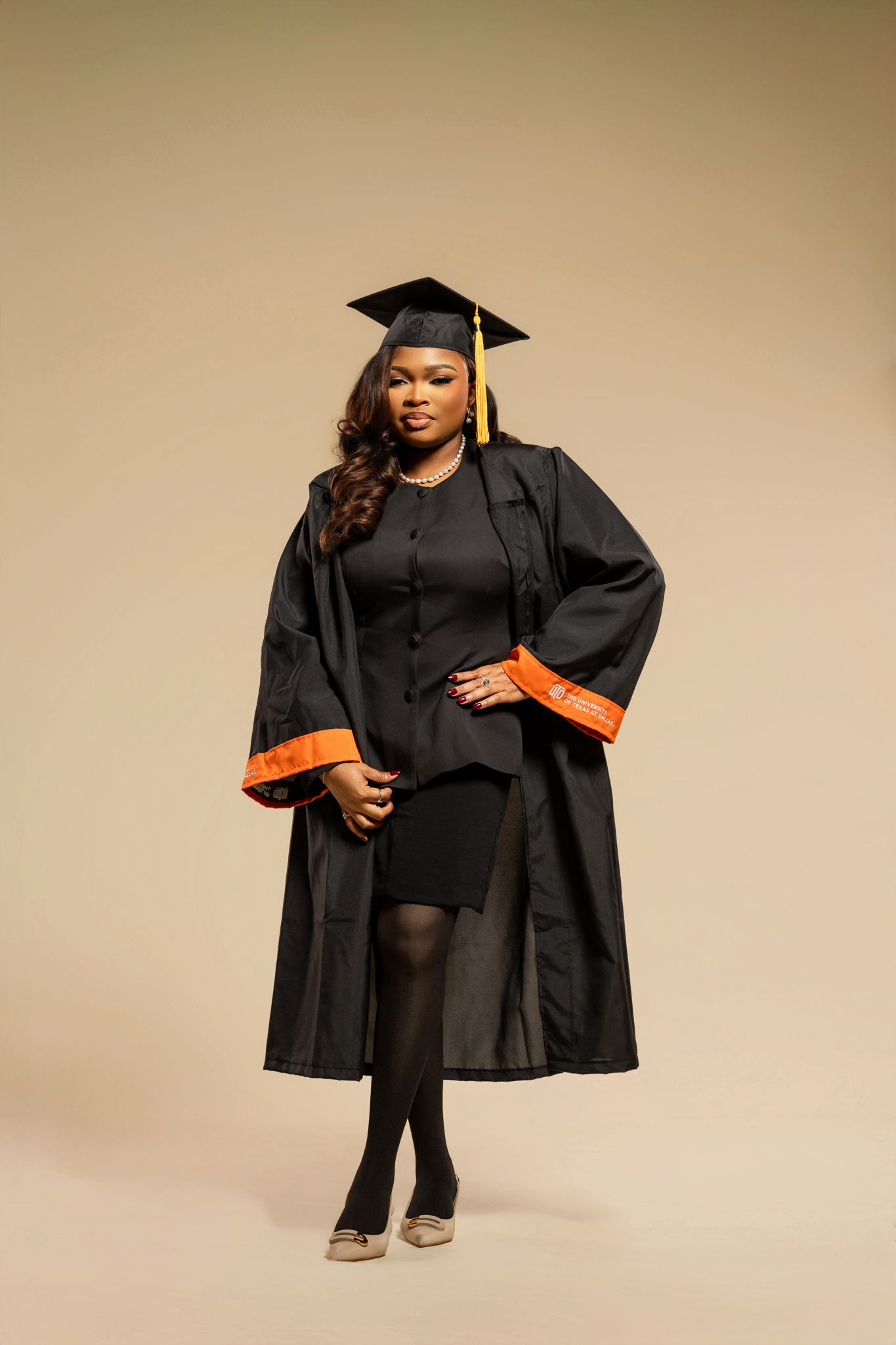 Graduate in black cap and gown poses confidently against beige background in professional graduation photoshoot series.
