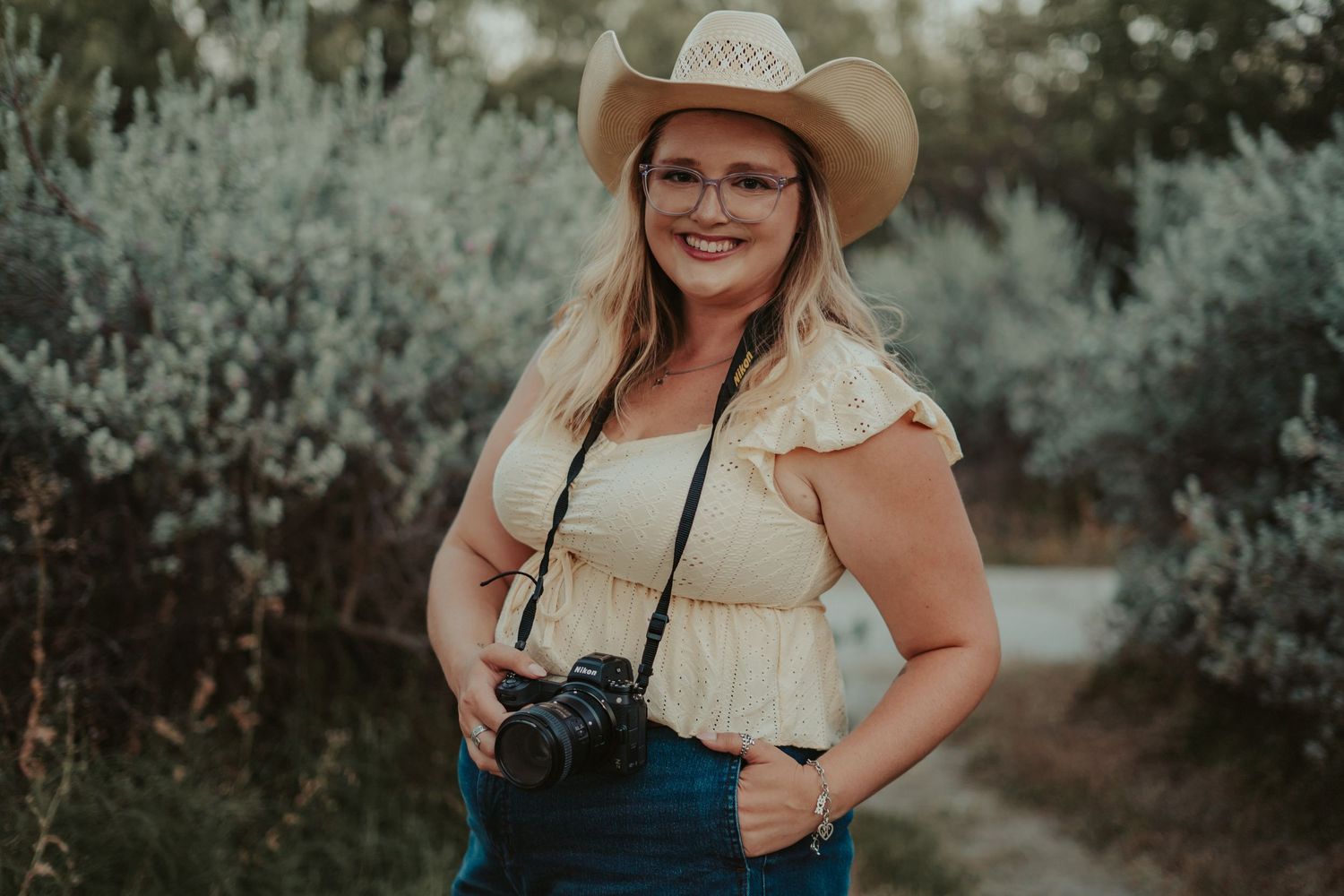 Smiling blonde woman in cowboy hat and glasses holding camera outdoors among green shrubs.