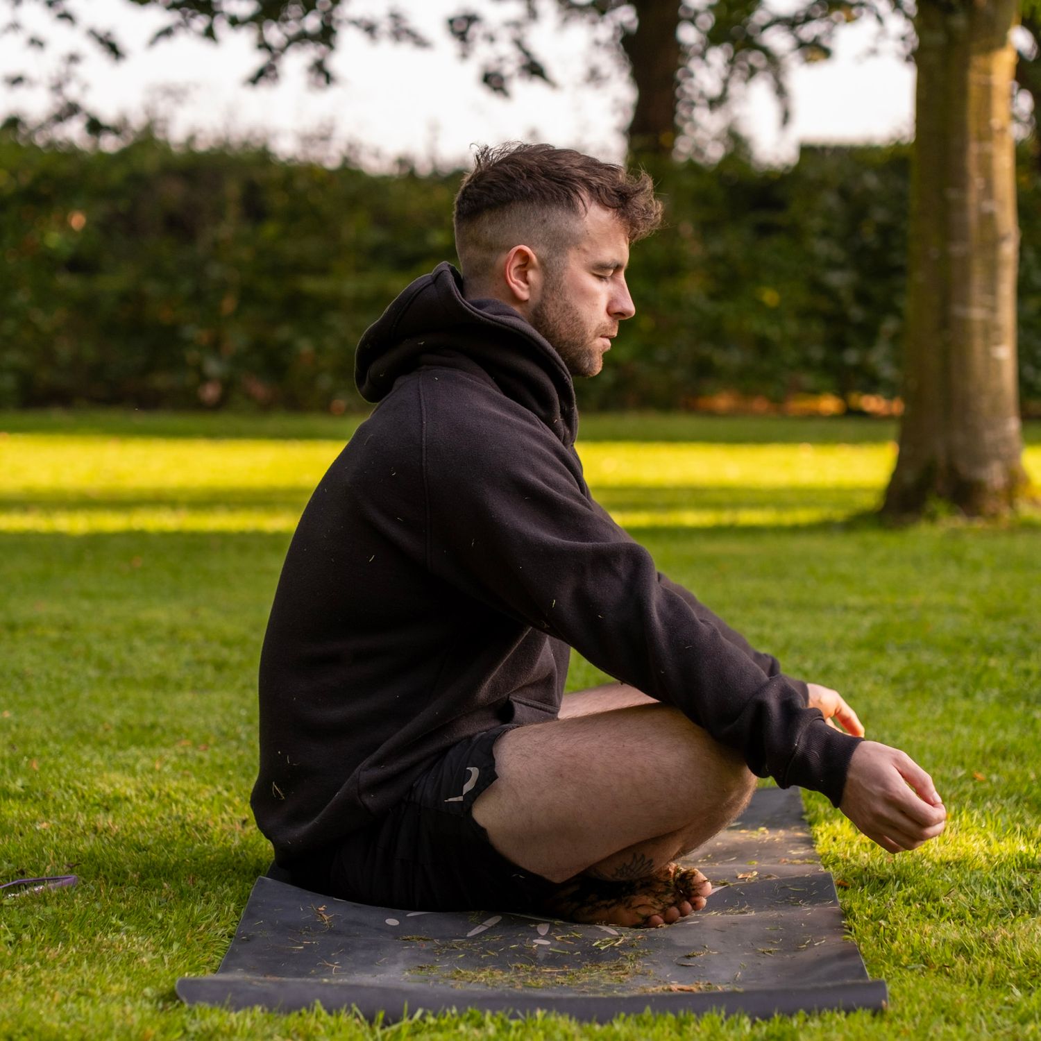 Person meditating on yoga mat outdoors in black hoodie against sunlit grass backdrop.