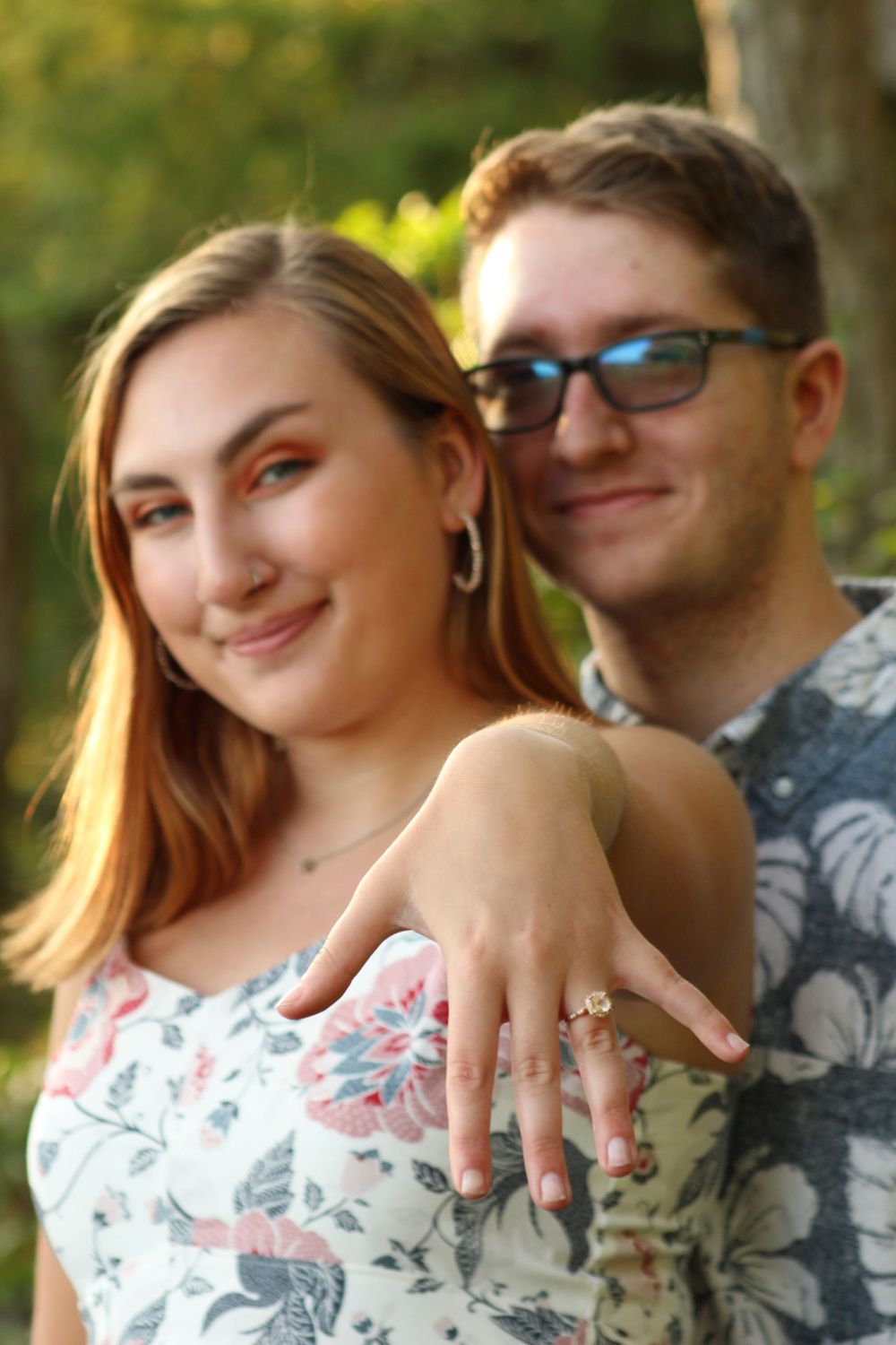 A newly engaged couple shows off an engagement ring while posing in floral patterned clothing.