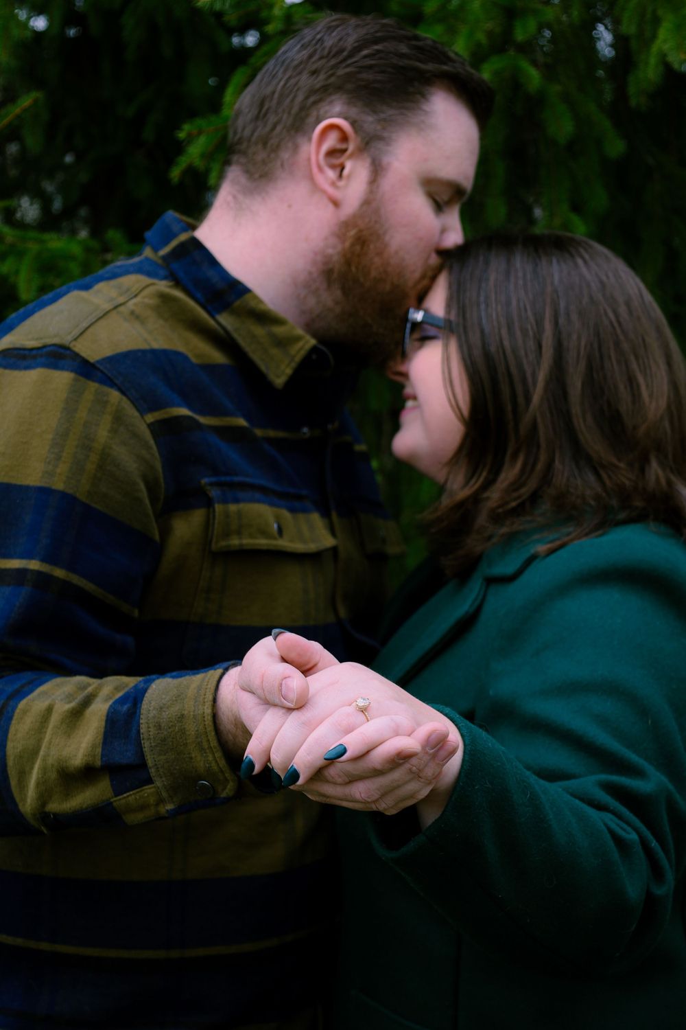 A romantic engagement moment shared between a couple in front of evergreen trees.