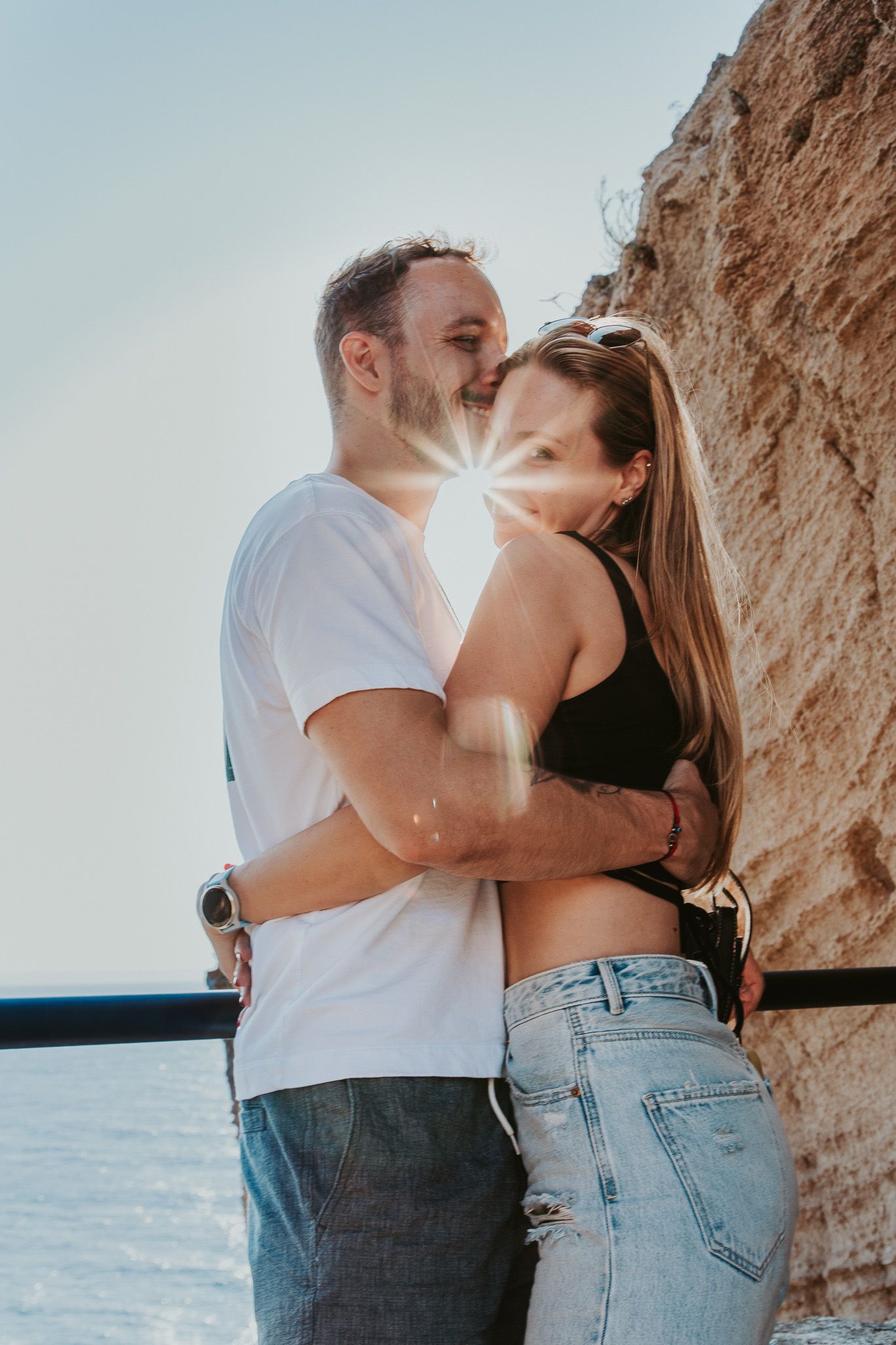 A couple embraces against a rocky cliff backdrop near the ocean  - love shoot at sunset