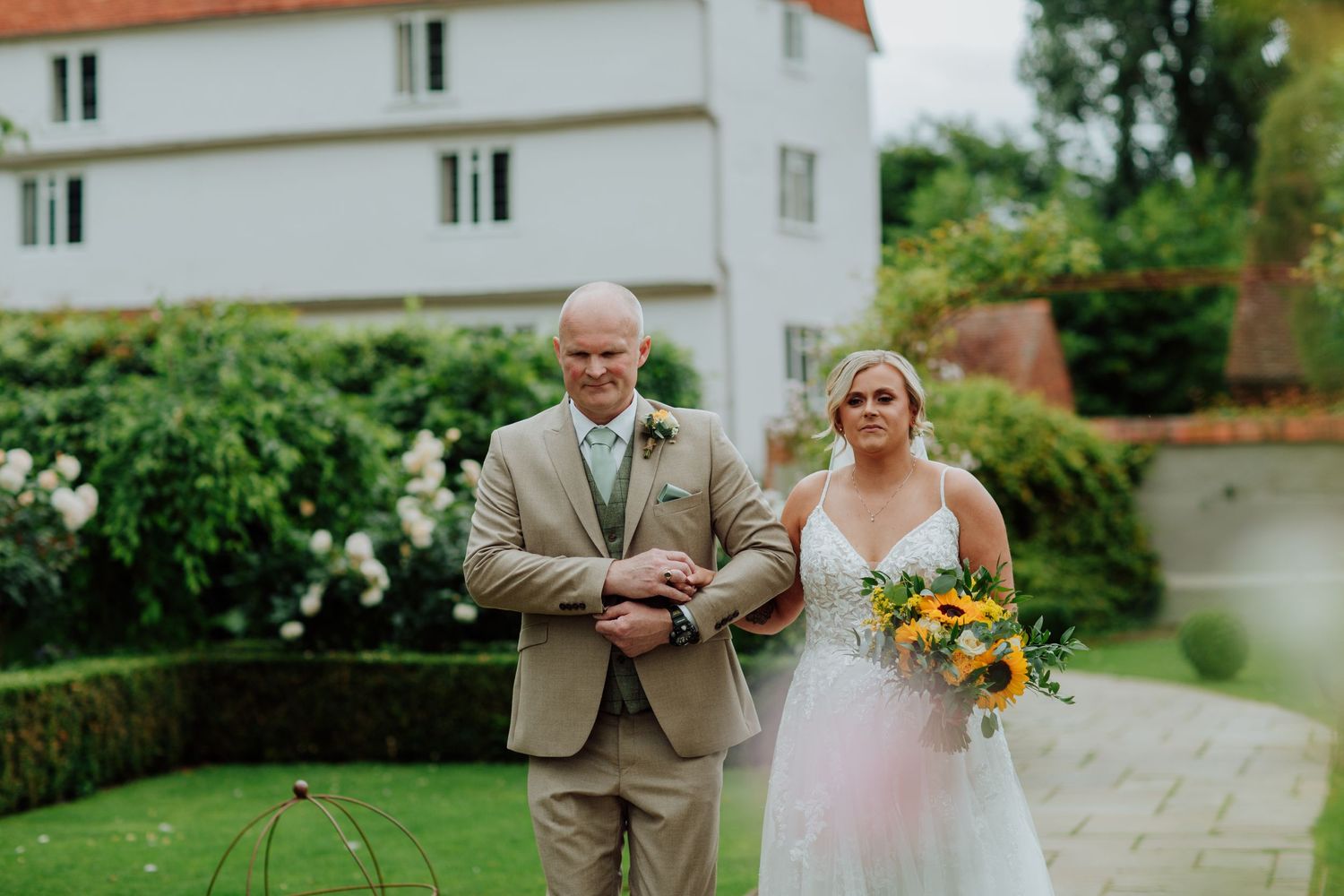 Couple stands together on garden path with white house in background. Father and daughter wedding ceremony Houchins