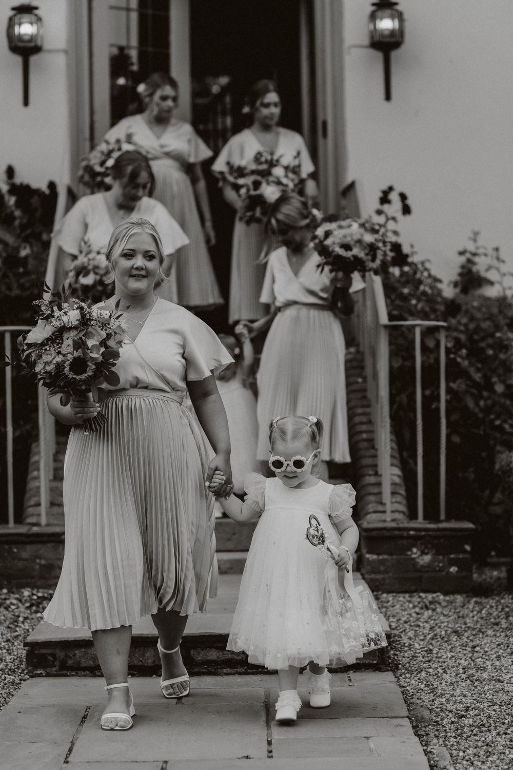 Wedding party members in light colored dresses walk down stone path carrying bouquets.