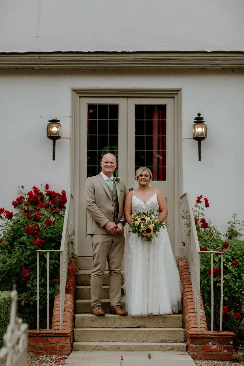 Wedding couple poses on front steps of white building with red flowers and lanterns. Houchins wedding venue