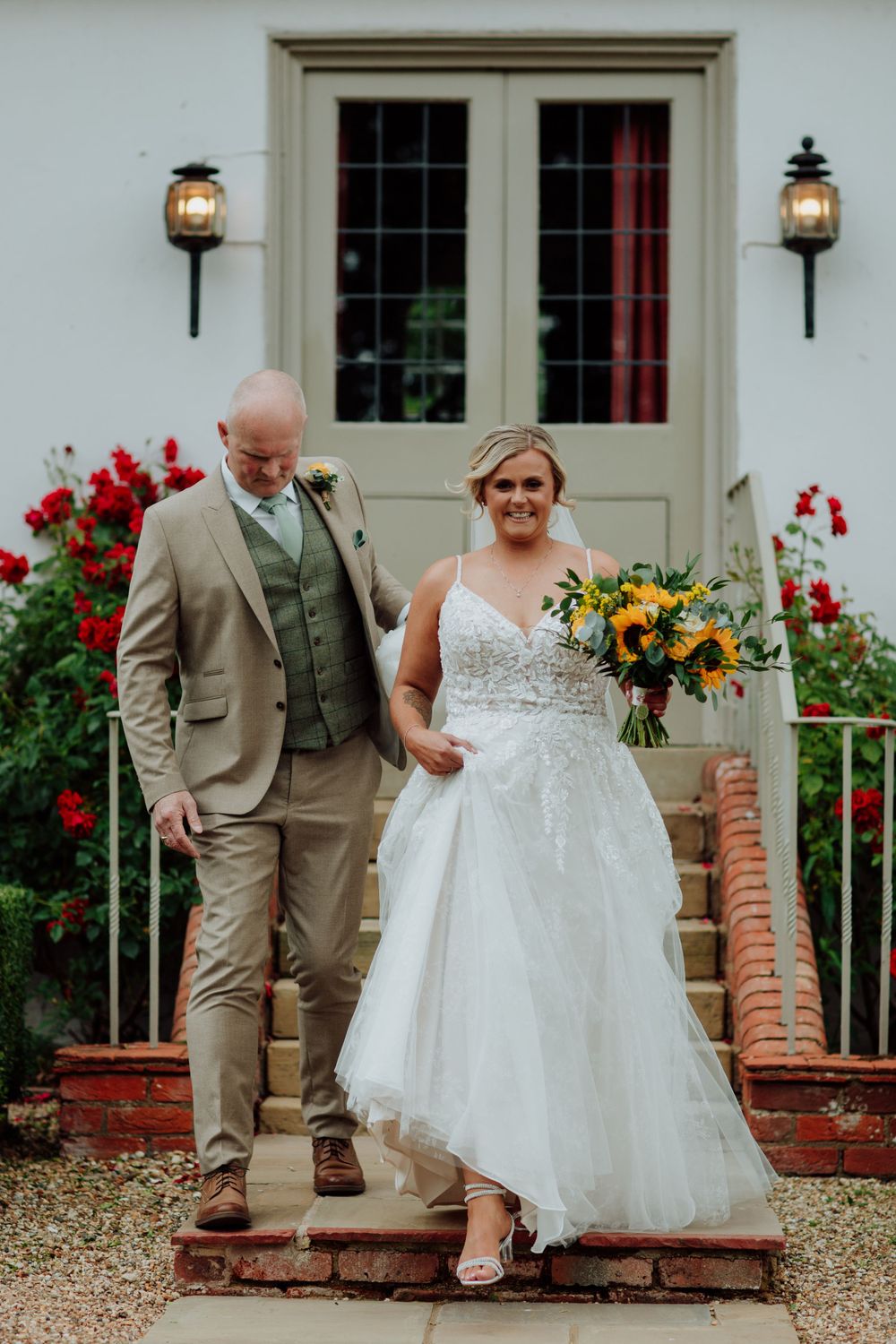 Wedding couple descending brick steps in front of white building with red flowers. Houchins Essex wedding venue