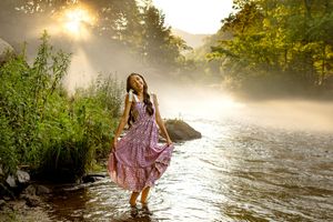A young Senior in a floral dress wades through a misty river at sunrise, surrounded by lush green trees in Bryson City, NC