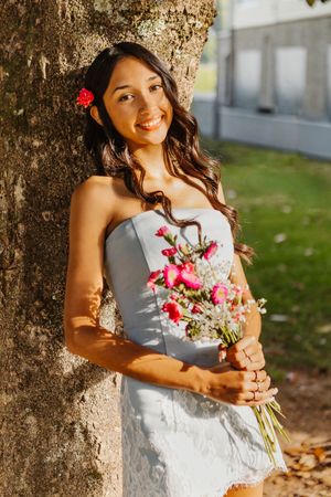 Young woman in white strapless dress holding pink flower bouquet, leaning against tree, smiling outdoors at Lake Junaluska, NC