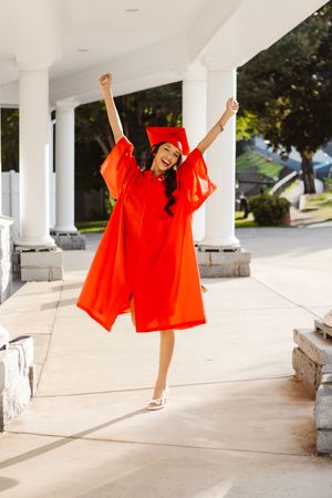 A joyful young woman in a red graduation cap and gown celebrates with arms raised outdoors near white columns near Asheville, NC