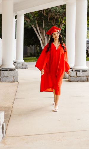 A smiling graduate walks in a red cap and gown between white columns on a sunny day in the Appalachian Mountains at Lake Junaluska