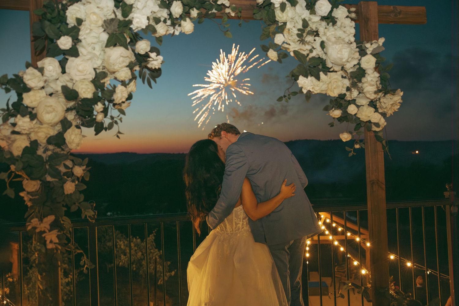 Romantic kiss under floral arch with fireworks and sunset backdrop.