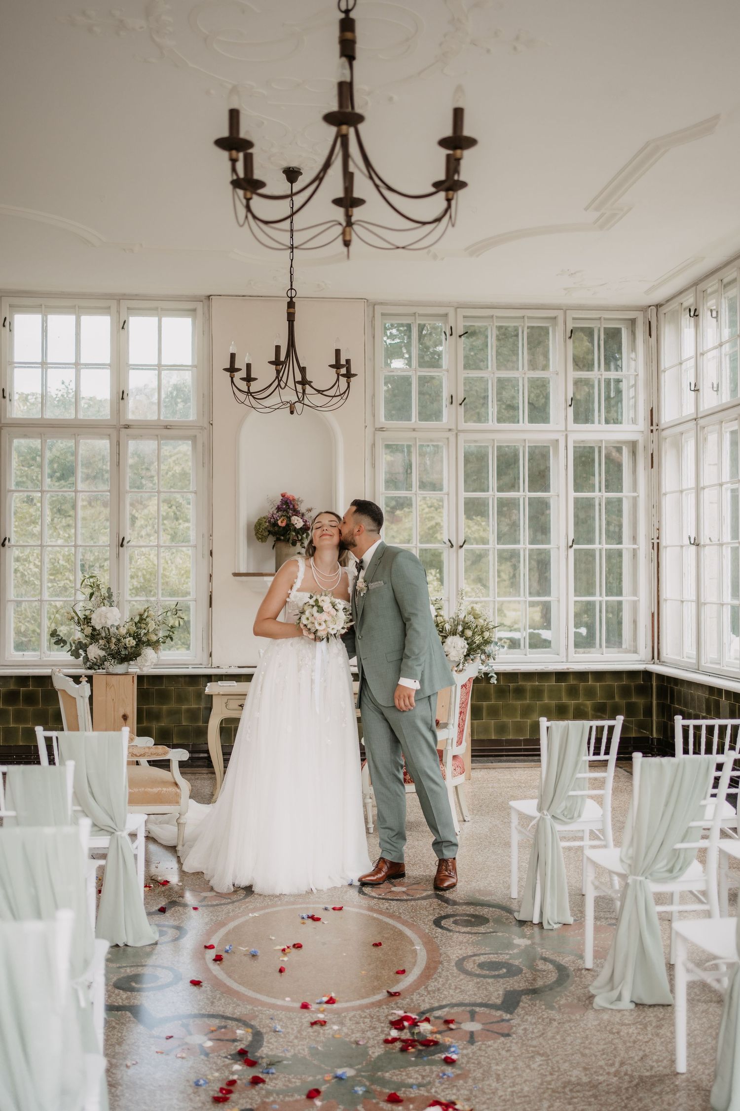 Wedding ceremony setup in bright conservatory with chandelier and rose petals on floor.