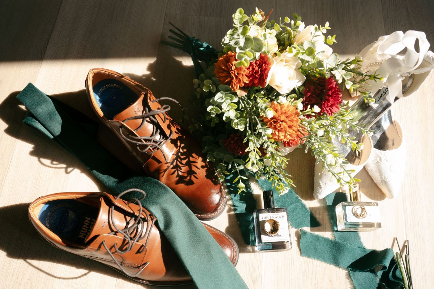 Brown leather oxford shoes, green tie, floral bouquet, and wedding accessories arranged on a wooden floor.
