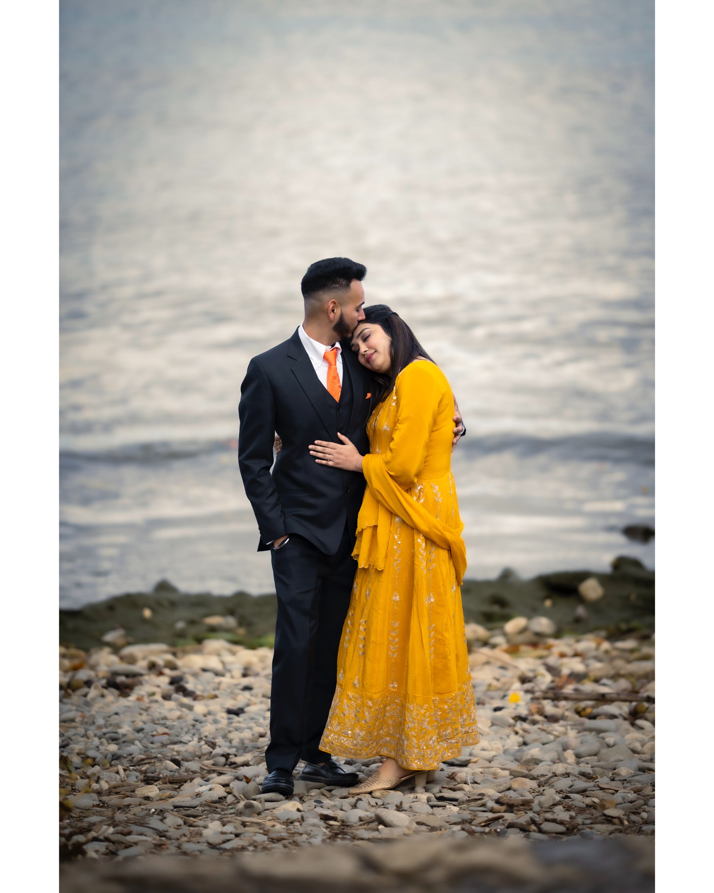 Couple embracing by rocky shoreline with bride in vibrant yellow dress against ocean backdrop.